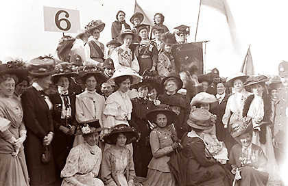 Foto av massor av suffragetter, många iförda stora hattar, utomhus. I bakgrunden står några suffragetter på vad som förmodlingen är en upphöjd talarstol. Fanor viftas med i bakgrunden. Längst till höger är syns hälften av en polisman som bevakar damerna