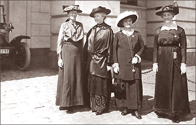 Foto av fyra damer i länga klänningar och hattar på gatan. I bakgrunden anas en bil