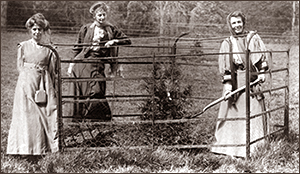 Foto av tre kvinnor, två utanför och en inne i en inhägnad. Hon inne, Emmeline Pethick-Lawrence, håller en spade och ett nyplanterat träd syns i mitten. Alla ser in i kameran och ser glada ut.