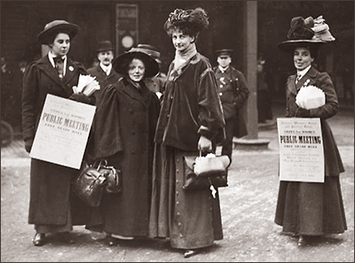 Foto av fyra kvinnor som står uppställda och tittar in i kameran. Både Mary Gawthorpe och Constance Lytton bär på väskor. De andra bär plakat om ett offentligt möte. I bakgrunden syns en man som ser ut att vara polis och en annan man.