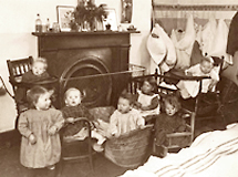 Foto av sju småbarn (1-2 år) som sitter i stolar eller står på golvet i daghemmet . I bakgrunden något som skulle kunna vara en eldstad och ännu längre bak hänger tygpåsar på hängare på väggen.