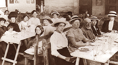 Foto många kvinnor och barn och någon man som sitter vid matbord som står på rad. Flera tittar in i kameran.