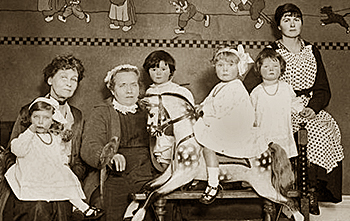 Foto av Emmeline och de fyra fattiga flickor som hon adopterade, alla flickorna i samma storlek, cirka 3-4 år. Med i bilden finns också en äldre kvinna med en papegoja samt en yngre kvinna med förkläde på sig. Den yngre ser ut som en barnsköterska. Flickan i förgrunden sitter på en trähäst.