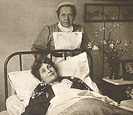 En sköterska står vid huvudänden av Emmelines säng som hon ligger i. Nattygsbord till vänster med blommor och ett fotografi på