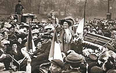 Foto av Emmeline som står i en öppen bil omgiven av folkmassor. Hon ser jublande glad ut och sträcker upp ena armen, i den andra håller hon blommor