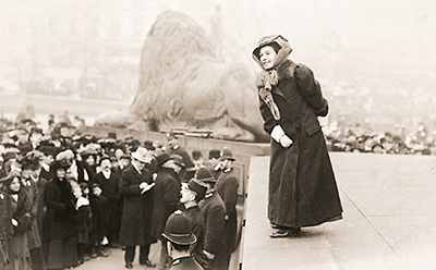 Foto av Emmeline som står högt ovanför en folkmassa och håller tal. Hon har hatt och sjal och tjock kappa på sig, och åskådarna är också väl påklädda. Längst fram, nedanför Emmeline, står en rad poliser och bakom henne, mitt i bild, är ett jättelikt marmorlejon sett bakifrån