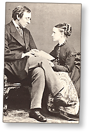 Foto av Henry Fawcett som sitter med några papper i knät och Millicent som sitter på golvet bredvid honom och allvarligt ser upp på honom.