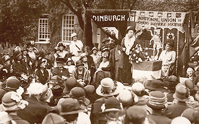 Foto av millicent Garrett Fawcett som håller tal utomhus för massor av kvinnor . En banderoll för NUWSS hänger längst fram liksom en för staden de befinner sig i, Första bokstaven har blåst och skyms, men det går att förstår attd et ska stå "Edinburgh". På talarstolen bredvid henne står ne massa blommor