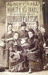 Foto av sex kvinnor vid mitten av 1800-talet framför en stor affisch med text om rösträttsmöte för kvinnor i Albert Hall samt en demonstration för rösträtt