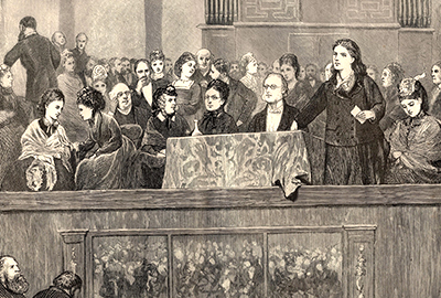 Rhoda Garrett håller tal stående vid ett bord med duk vid ett möte 1872  omgiven av andra dåtida feminister som sitter runt omkring henne