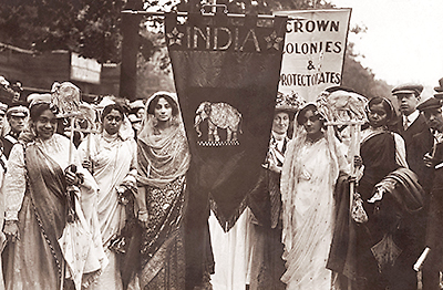 Foto av en stor grupp indiska kvinnor, i indiska kläder och sjalar på gatan i ett demonstrationståg. De mittersta bär ett standar som det står "India" på och en bild av elefant.