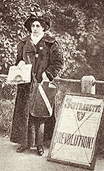 Foto av Sophia som står och säljer  WSPU:s tidning The Suffragette. Bredvid henne står en gatuskylt med  tidningens logotype och med stora bokstäver skrivet: REVOLUTION!