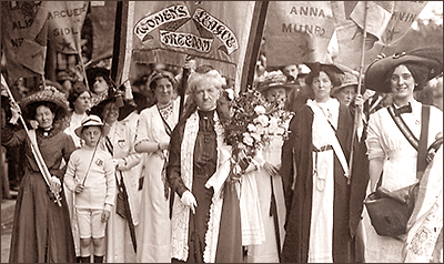 Foto av demonstrationståg med Charlotte Despard längst fram. Hon bär en blomsterkvast. Bakom henne syns ett tjusigt sytt standar med texten "Women's Freedom League". Flera demonstranter ser glada ut, men hon är allvarlig.