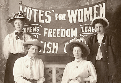 Foto av fyra WFL-are, två sittande och två stående med en banderoll bakom med texten "Votes for Women - Women's Freedom League" på och något mer oläsligt  underst