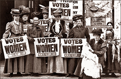 Foto av ett antal kvinnor som håller skyltar med texten "Votes for Women" och WFL på. En kvinna sitter ner med en baby i famnen.