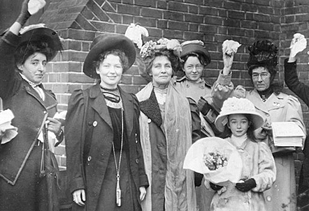 Fem kvinnor och en liten tjej med stor hatt och blommor i handen. Alla ler och några lyfter knutna nävar i luften. Christabel och Emmeline Pankhurst i mitten