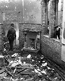 Foto av en polis som går omkring i ruinen av ett hus, medan andra poliser tittar in genom öppningarna i en murad vägg där fönster funnits