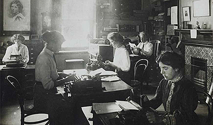 Fyra kvinnor sitter vid olika skrivbord och skriver på skrivmaskiner, ne man sitter i högra hördet och läser en tidning, ljus strömmar in från ett fönster mitt i bilden. Bredvid fönstret finns en tavla föreställande Christabel Pankhurst.