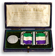 Uppfällt, mörkt färgat etui med en medalj som har suffragetternas färger på ordensbandet. I locket syns guldtext på ljusgul botten, men svårläst, det som syns är namnet Nellie Hall.