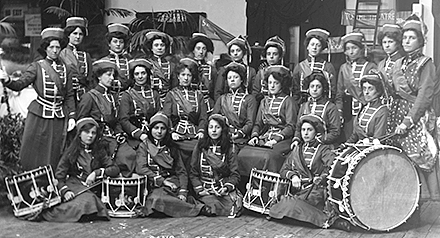 22 kvinnor i uniformer och hattar. De står med sina musikinstrumt, i förgrunden trummor i olika storlekar.