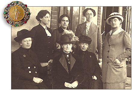 Foto av sju rösträttskvinnor med hattar och kappor, fyra stående och tre sittande. I vänstra hörnet över fotot finns en rösträttsbrosch