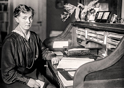 Foto av Eva Andén framför sitt skrivbord. Hon är klädd i en sidenblus med  länga manschetter mned klädda knappar och hon ser rakt in i kameran