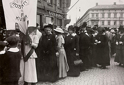 Foto av demontsration, det framgår att det är i Göteborg av en affärsskylt och av deras standar. Kvinnor i långa klänningar och stora hattar står i rad.