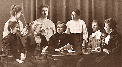 Foto som ser ut att vara ett montage. Åtta kvinnor sitter respektive står runt ett bord, alla sitter riktade mot kameran. Frigga Carlberg i mitten tittar på sin bordsgranne till vänster.