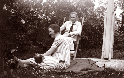 Foto av Nelly Thüring som sitter på marken framför Agda Östlund som sitter i en solstol i en trädgård. Båda ser glada ut, Melly ler stort, där hon sitter i profil. Till höger syns nederdelen/fästet till en flaggstång, bakom dem är en häck av buskar