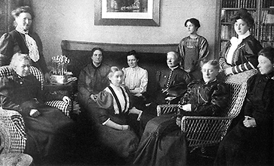 Foto av tio kvinnor i sekelskifteskläder som sitter i ett vardagsrum. I bakgrunden skymtar en tavla och ett bokskåp med glasdörrar. Alla ser allvarliga ut och de flesta tittar mot kameran