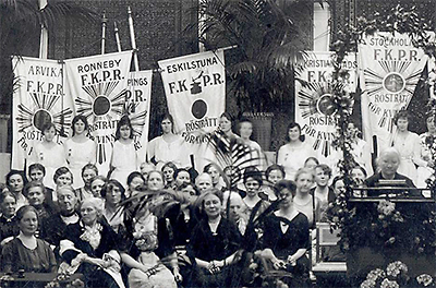 Foto av rösträttsmöte med massor av kvinnor och standar för olika städers FKPR. Längst till höger står Ellen Key i en talarstol och håller tal.