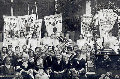 Foto från möte med LKPR. I bakgrunden standar med texten F.K.P.R. och olika ortsnamn på olika standar: Arvika, Ronneby, Eskilstuna, Kristianstad och Stockholm. Unga kvinnor i vita klänningar håller i standaren. Framför dem sitter en mängd kvinnor och till höger står en vithårig Ellen Key i en talarstol. Längst bak syns stora krukor med palmer