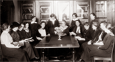 Foto av femton kvinnor som sitter och står runt ett bord. Mitt på bordet står en vas med blommor. I bakgrunden böcker i bokskåp som det står "Skönlitteratur" på.