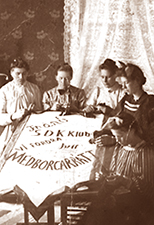 Foto av fyra kvinnor som sitter och syr på ett standar. I bakgrunden en gardin och tapet. På standaret syns texten Frånö och "Vi fordra medbergarrätt" och något mer svårläst
