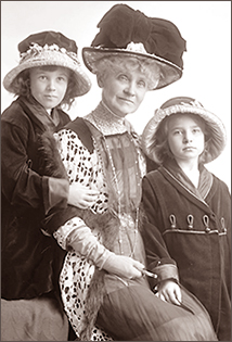 Foto av en äldre kvinna som sitter omgiven av två små flickor. Alla tre har stiliga hattar på sig