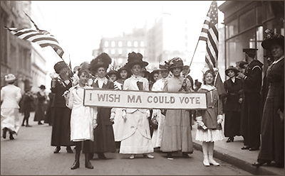 Ett antal kvinnor och barn med amerikanska flaggir syns i en parad på gatan. Barnen längst fram bär ett liggande standar med texten: I Wish Ma Could Vote"