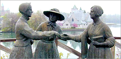 Foto av del av en staty föreställande två kvinnor som räcker fram handen och en tredje som ser ut att ha presenterat dem för varandra. I vakgrunden en disig byggnad