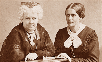 Foto i halvfigur av Elizabeth Cady Stanton och Susan B. Anthony vid ett bord som anas vid deras händer. Elizabeth ser rakt in i kameran, medan Susan tittar snett åt sidan.