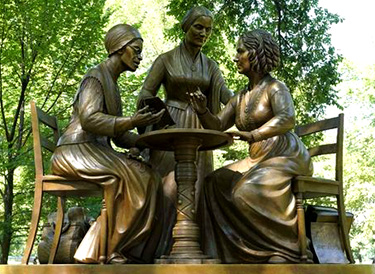 Foto av en staty med rvå kvinnor som sitter på var sida om ett litet runt bord, mellan dem står en kvinna. Det är som om de är mitt uppe i diskussioner