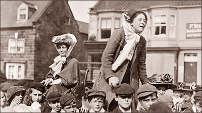 Foto av Margret Bondfield i yngre dar, hon står på något som gör att hon syns högt över alla huvuden och hon håller tal. I bakgrunden syns Emmeline Pankhurst, som också finns över alla huvuden. Omkring dem är folk på en gata med hus i bakgrunden