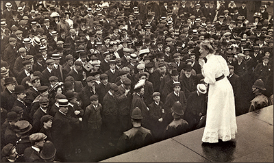 Foto av Mary Macarthur i ljus klänning där hon står på ett podie med massor av människor som lyssnar nedanför. Längst fram mot henne står en rad poliser och ser ut över folksamlingen