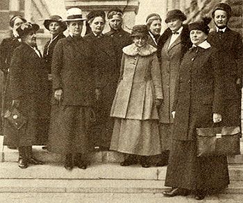 Foto av en grupp kvinnor som står utomhus på en trappa iförda kappor och hattar. Bertha Wellin står längst fram, några trappsteg nedanför, och håller en väska/portfölj i handen