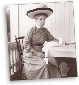 Foto av Gerda Meyerson som sitter på en trästol vid ett bord på vad som ser ut att vara en veranda till ett sommarhus. Hon har en stor hatt på huvudet och ser in i kameran
