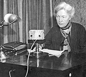 Foto av äldre Kerstin Hesselgren som sitter vid ett skrivbord och talar in ett föredrag för radio. Hon har papper framför sig och ser allvarlig ut.