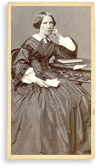 Foto av Rosalie som sitter vid ett bord och lutar armen mot en hög med böcker. I andra handen håller hon några papper, kanske brev