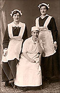 Foto av två pigor i uniform som står bakom en kock som sitter. Alla tre kvinnorna ser in i kameran.