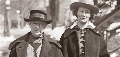 Porträttbild på två kvinnor som går på en gata, bägge iförda kappa och hatt