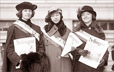 Foto av tre unga kvinnor i halvfigur som står med kappor och hattar på gatan och håller i tidningar The Suffragist. De har också band över brösten med cu:s färger gult, vitt och lila, även om det inte syns på en bruntonad bild