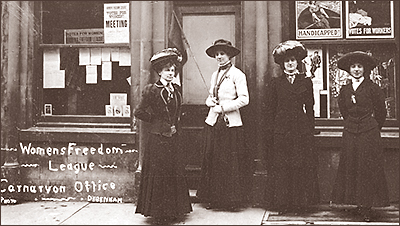 Foto av fyra kvinnor utanför WFL:s kontor i Debenham. Skyltfönstren är fulla av affischer. Kvinnorna har långa kappor och hattar på sig. Alla ser in i kameran