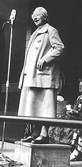 Foto av Anita Augspurd stående på ett podium och talande i en mikrofon. I bakgrunden sitter några kvinnor och lyssnar